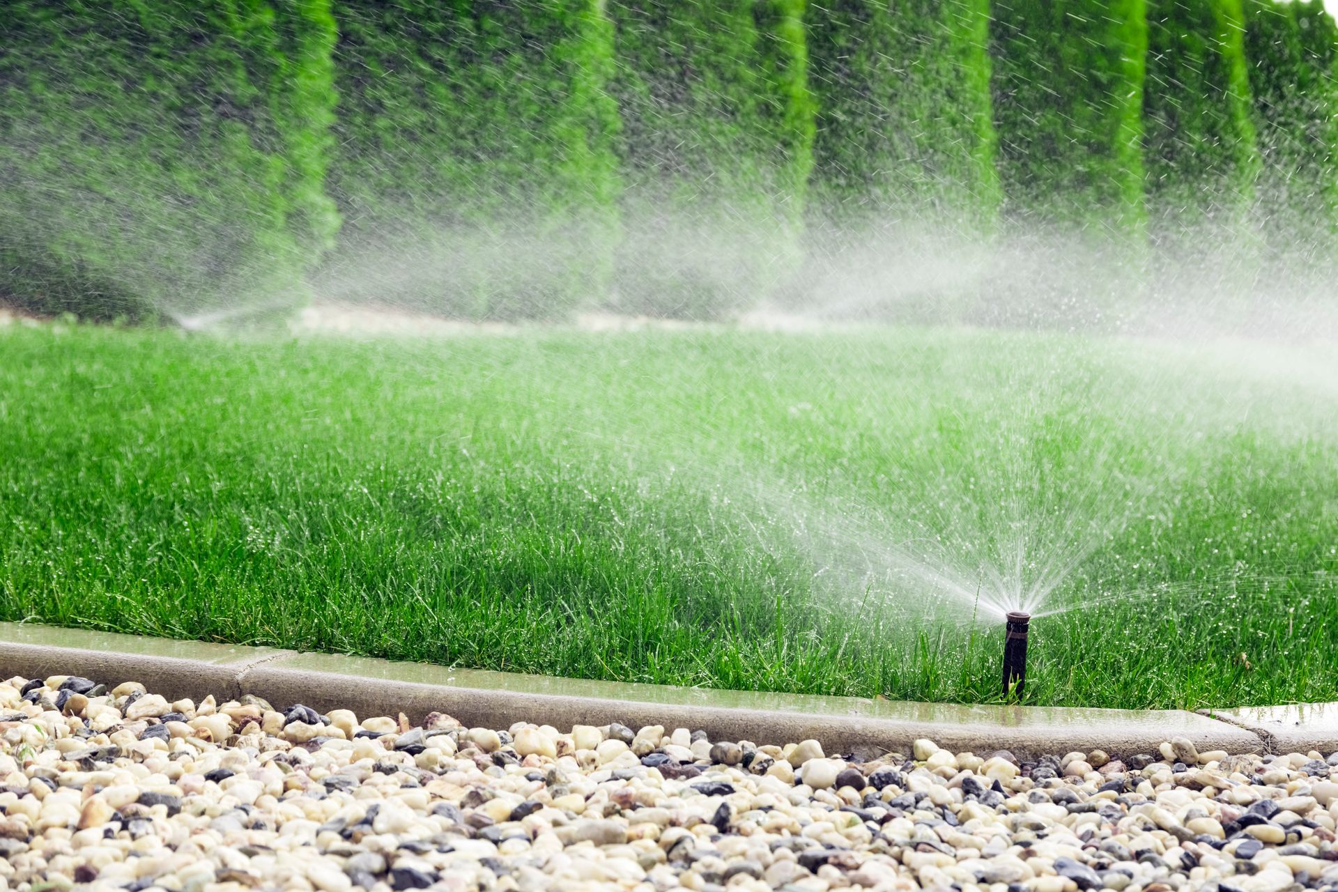 Lawn sprinkler spraying water across a green grass field beside a stone border