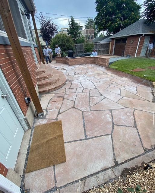Stone patio and walkway beside a brick house, with steps, retaining wall, and backyard seating area.
